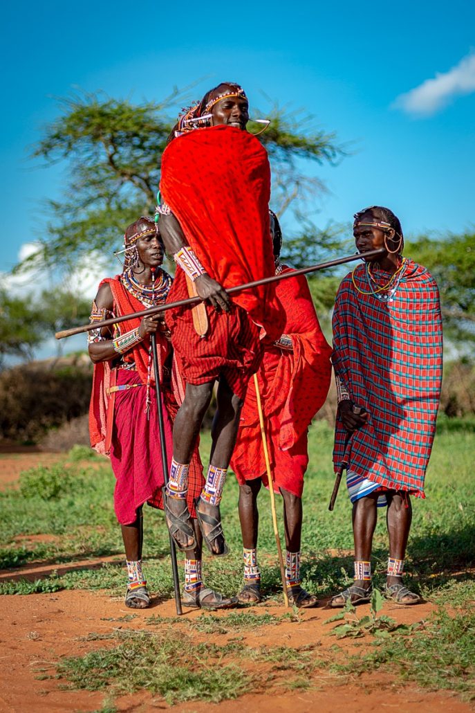 Portrait of a traditionally dressed jumping Masai warrior with stick in front of three Masai warriors.