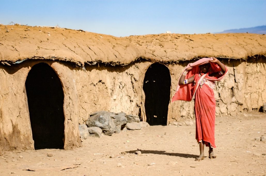 Portrait of a young Masai girl in traditional red robe in front of brown mud huts.