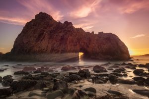 Photograph of a cliff in the sea with a hole through which sunrays fall.
