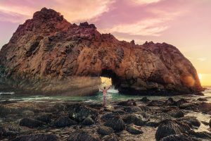 Photograph of a man in swimming trunks in front of a cliff in the sea with a hole through which sunrays fall.