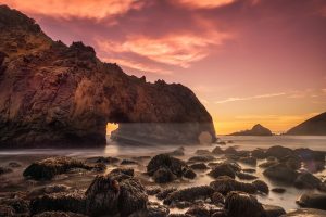Photograph of a cliff in the sea with a hole through which sunrays fall.