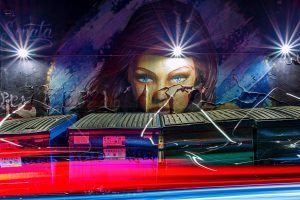 Night photograph of a woman's face painted on the wall behind garbage cans.