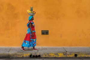Portrait of a fruit seller in traditional clothing with a fruit bowl on her head in front of a yellow wall.
