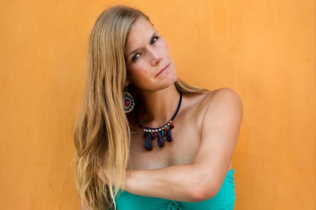 Portrait of a blonde young woman with a turquoise top in front of a yellow wall.