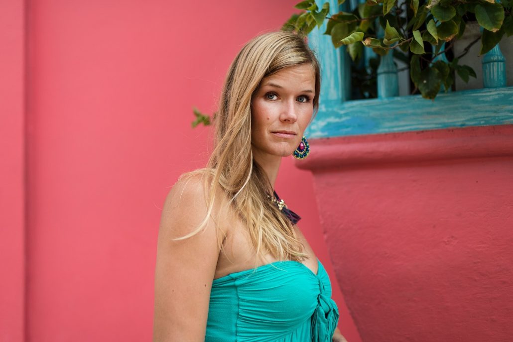 Portrait of a blonde young woman with a turquoise top in front of a pink house wall.