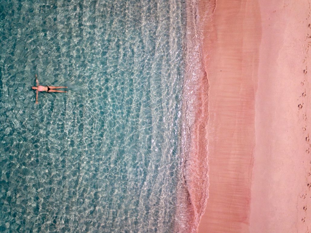 Aerial view of a woman swimming in the sea in front of a pink beach.