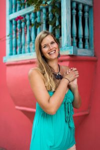 Portrait of a happy, blonde young woman with a turquoise top in front of a pink house wall.