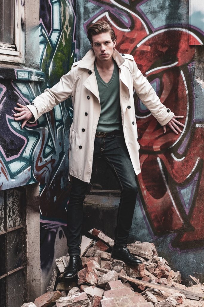 Portrait of a male model in a trench coat in front of a sprayed house wall.